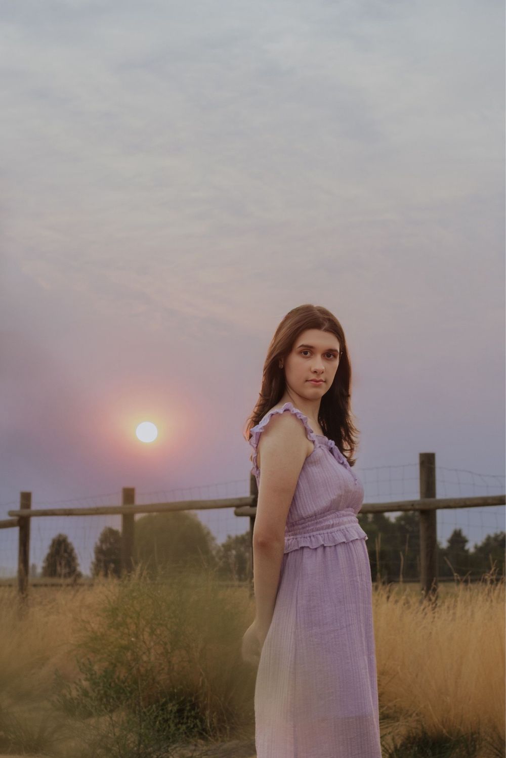 Girl standing in a lavender dress with hands behind back. Posed at sunset in Cheney, WA with sunlight leak.