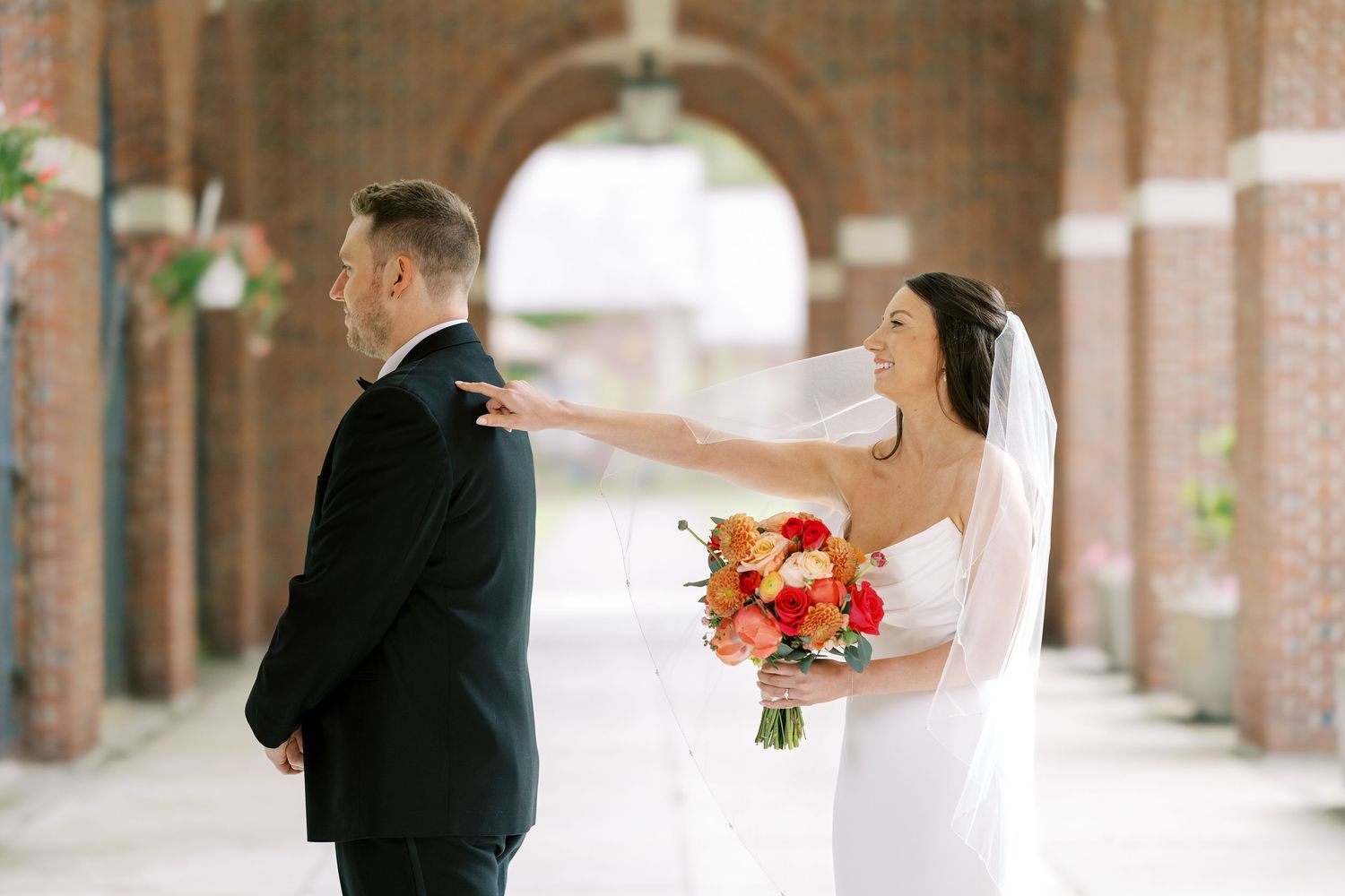 Couple in wedding attire stand under arched brick corridor while bride taps groom's shoulder holding pink bouquet at the Hall of Springs.
