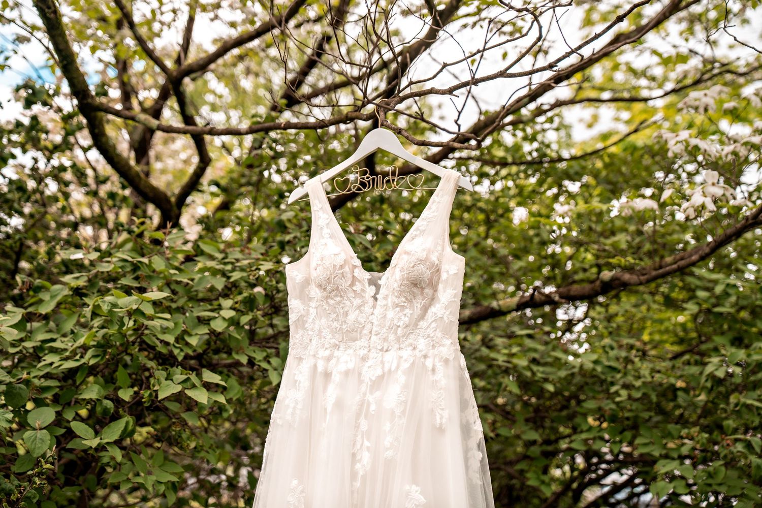 A white lace wedding dress hangs from a tree branch surrounded by spring foliage and blooming flowers.