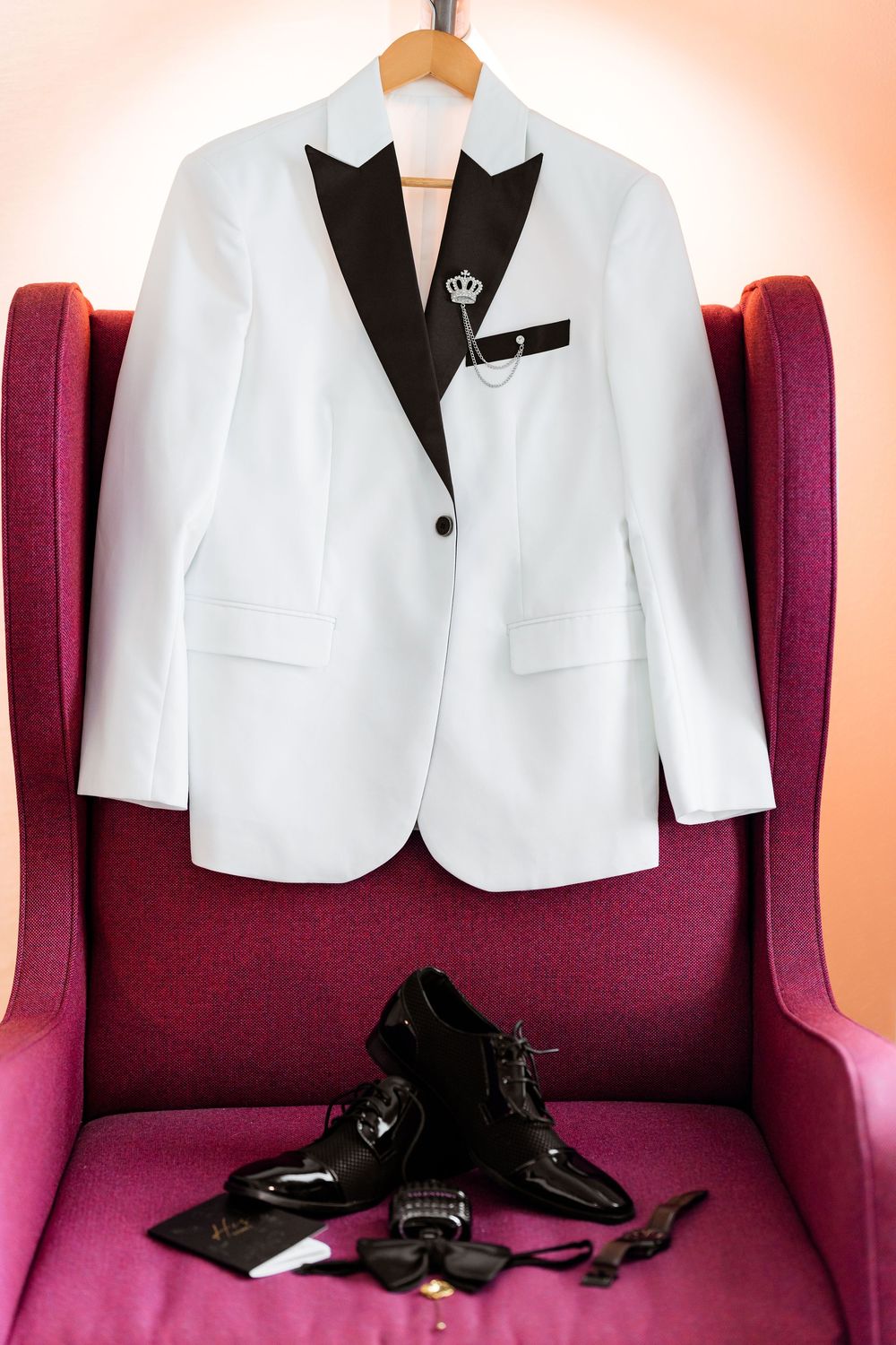 White tuxedo jacket with black lapels displayed on burgundy velvet chair with black dress shoes and accessories below.