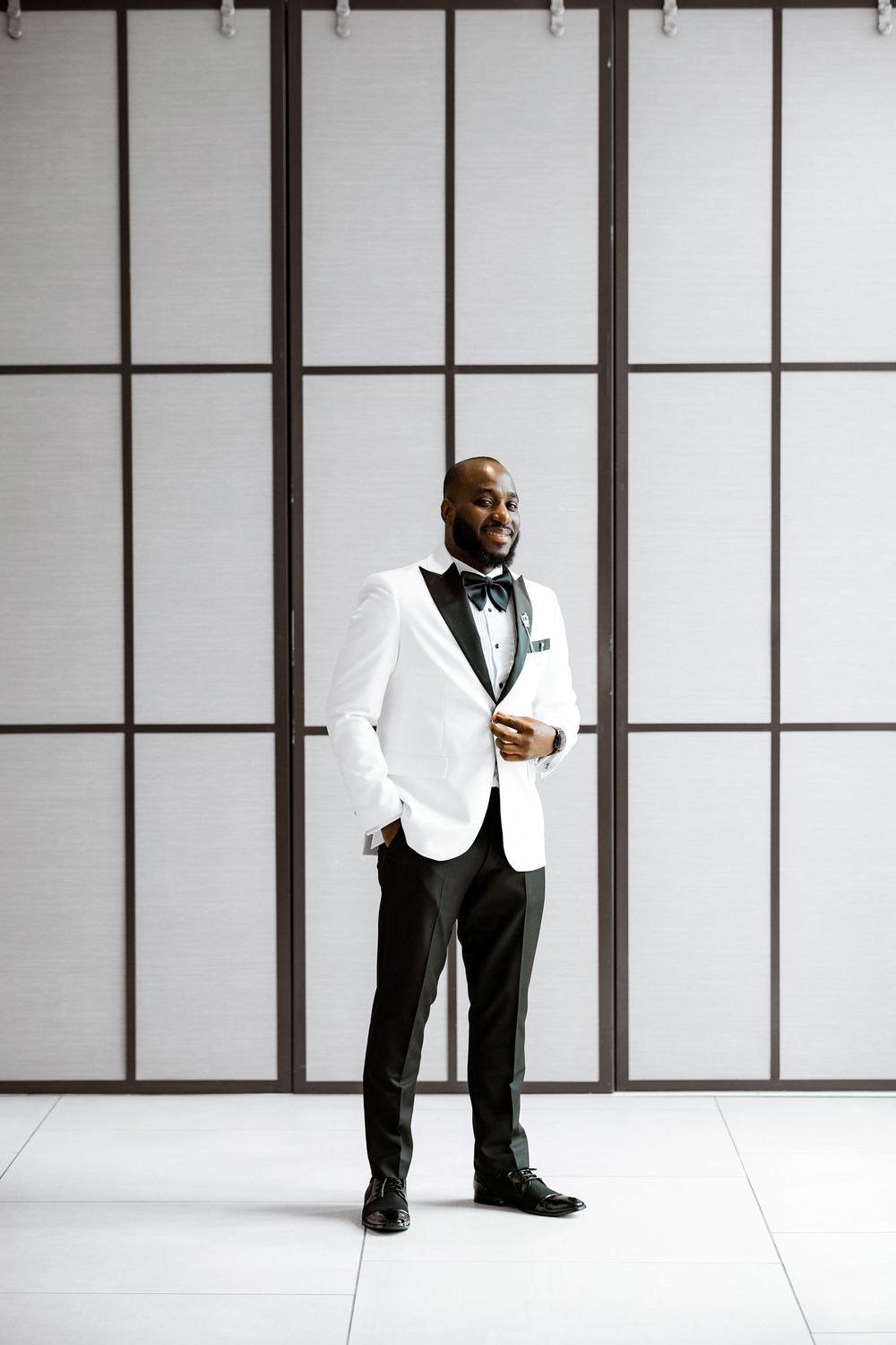 A person in a crisp white tuxedo jacket and black bow tie stands elegantly against a geometric paneled wall background.