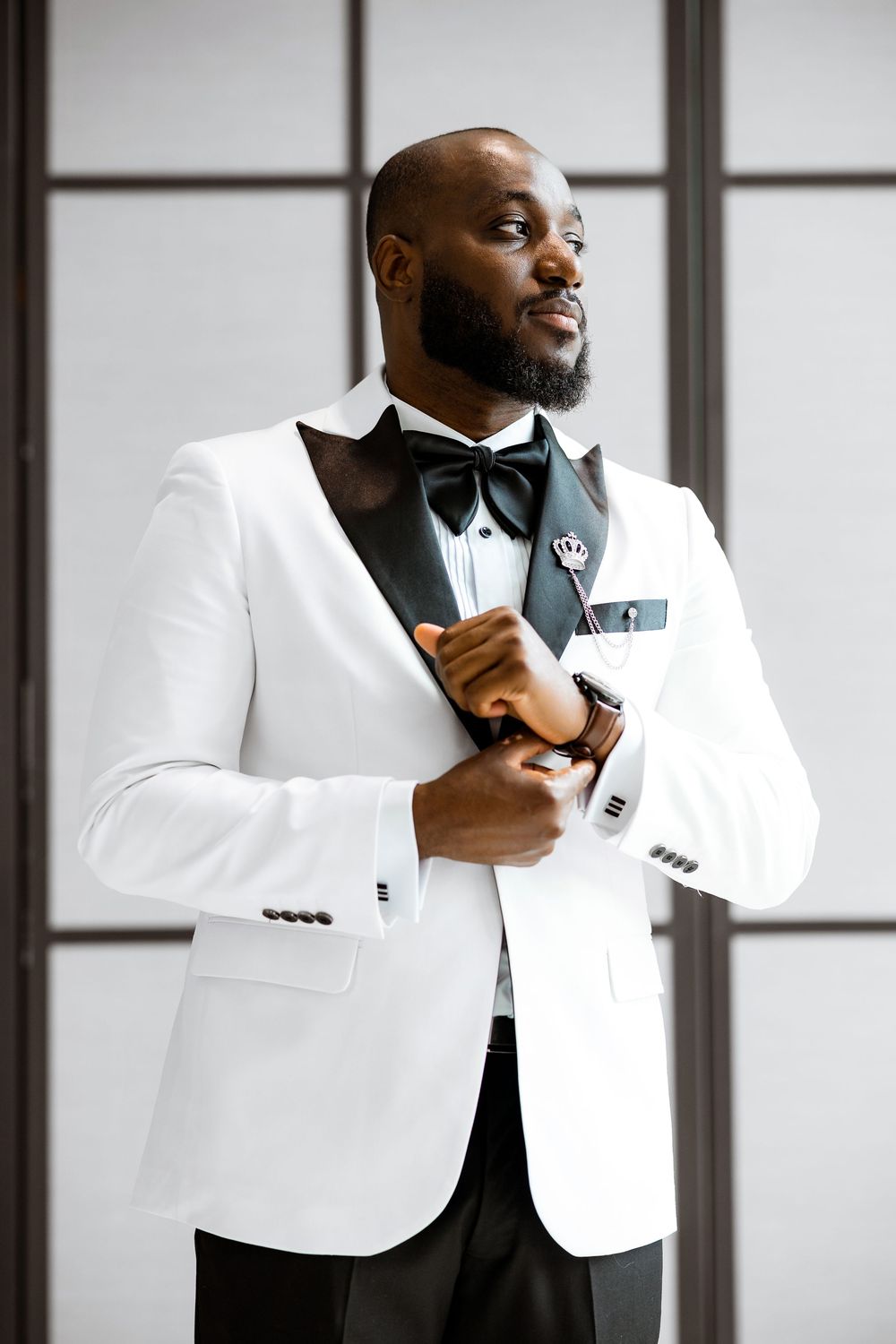 A stylish man in a white dinner jacket and black bow tie adjusts his shirt cuff while standing against a window backdrop.