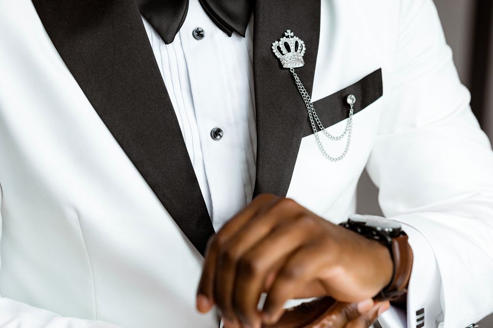 A close-up view of hands with a silver watch against a white and black tuxedo with a crown pin on the lapel.
