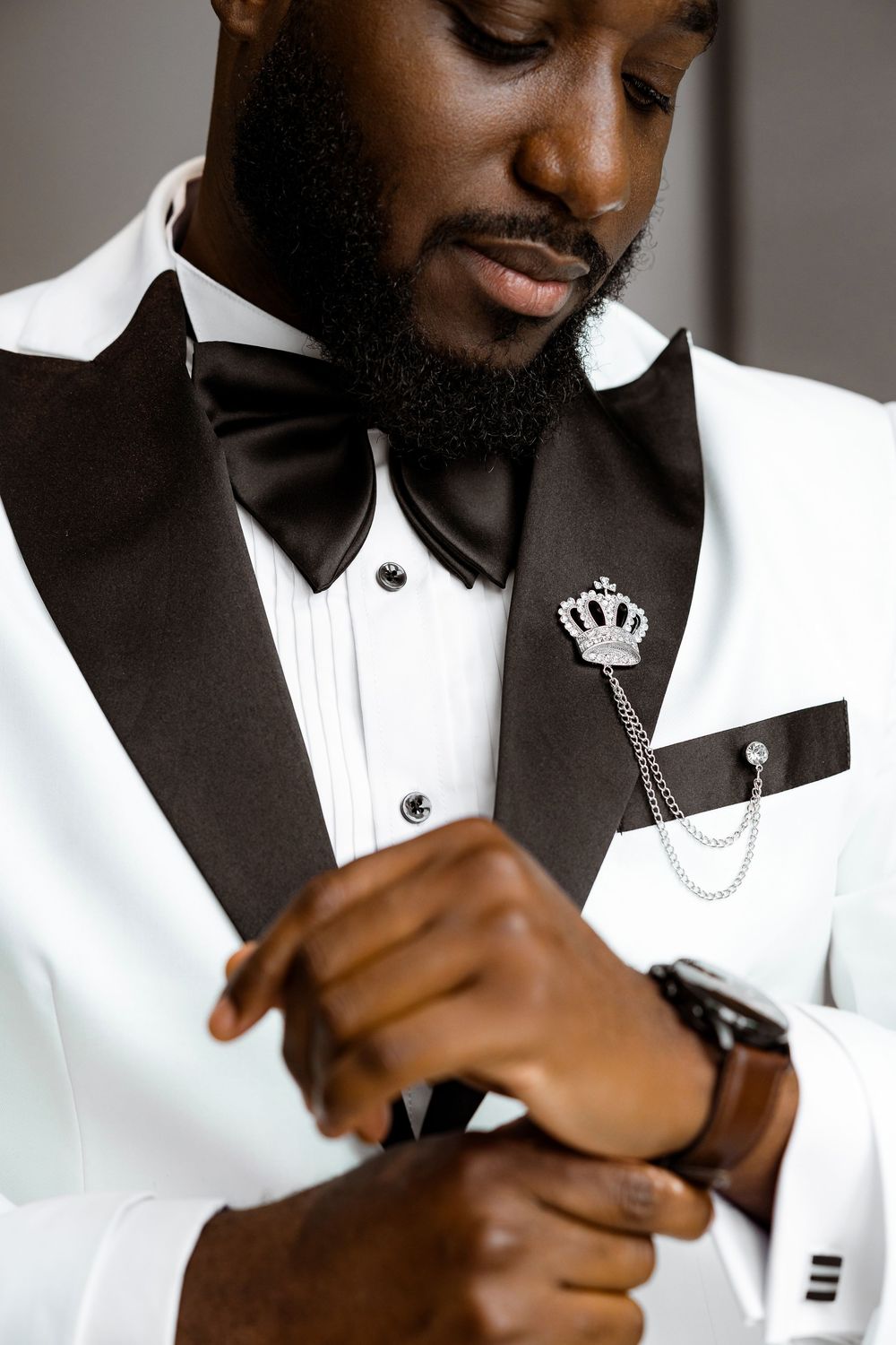 Elegant white and black tuxedo with crown brooch and chain accessory while adjusting watch.