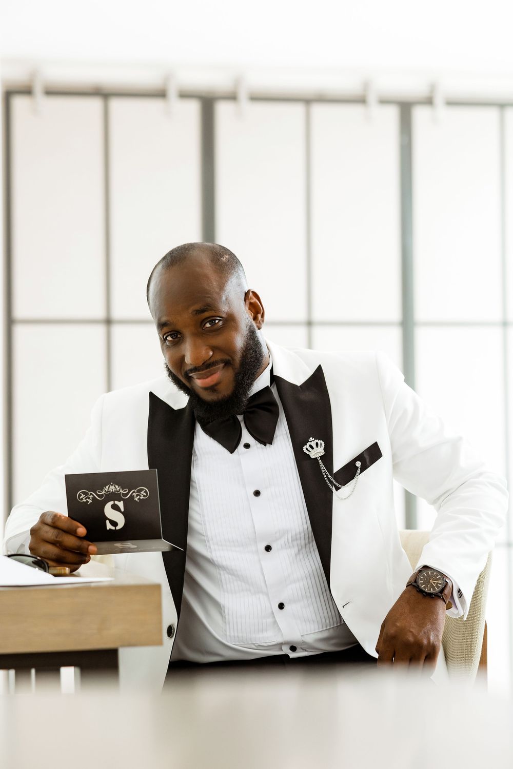 A well-dressed person in a white dinner jacket and black bow tie holds a small black card while posing at a table.