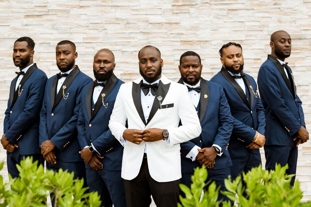 A formal wedding party wearing navy blue suits and one white tuxedo jacket pose together against a brick wall.