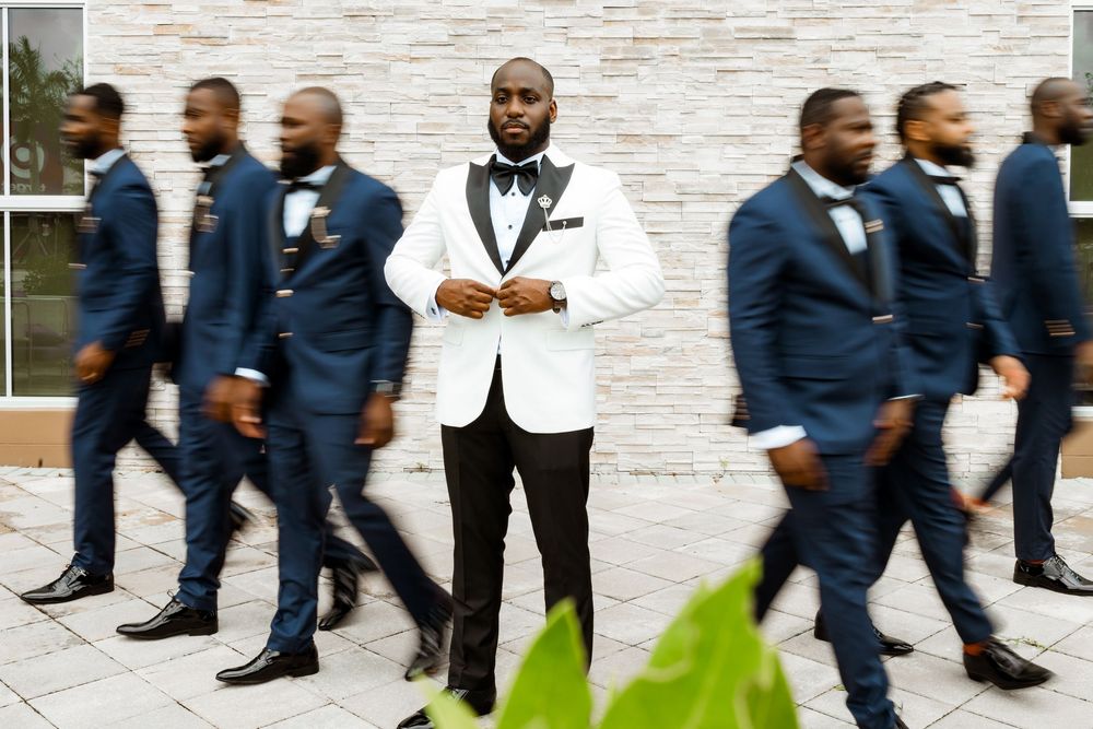 A groom in a white jacket stands centered while groomsmen in navy suits walk past against a light stone wall.