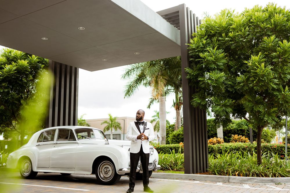 White vintage luxury car parked under modern building entrance with tropical palm trees and greenery.