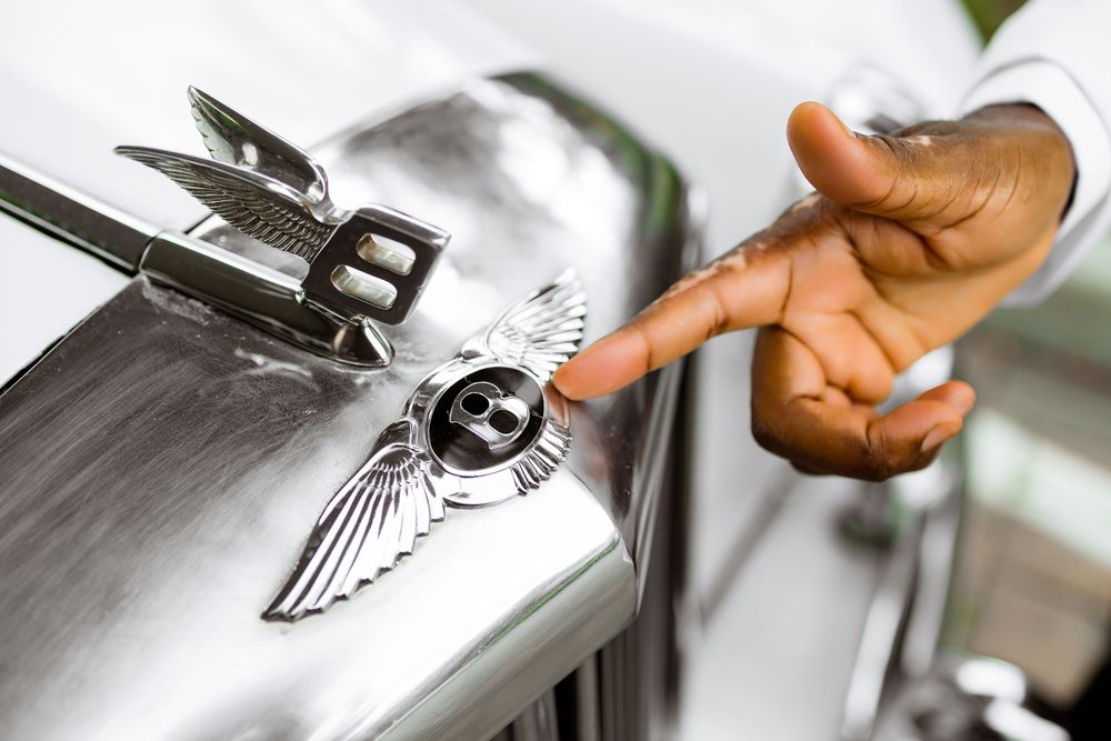 Close-up view of a hand touching the iconic Bentley emblem on a luxury car's chrome hood.