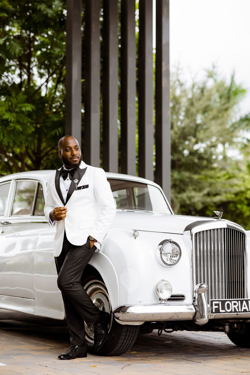 Classic white Rolls-Royce luxury car parked alongside stylish figure in white tuxedo jacket and black bow tie.