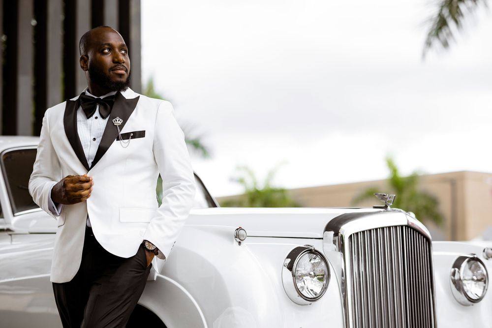 Elegantly dressed person in white tuxedo jacket standing beside vintage white Rolls Royce at outdoor venue.