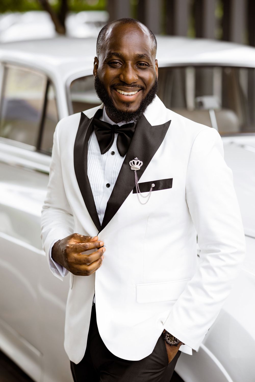 A well-dressed person in a crisp white tuxedo jacket with black lapels and bow tie smiles broadly beside a classic car.