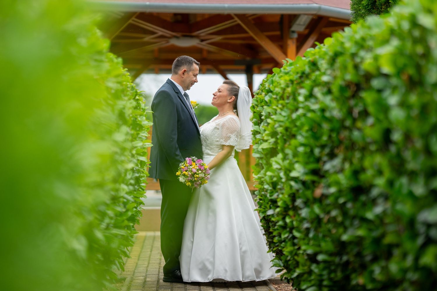 Couple shares an intimate wedding moment framed by lush green hedges under a wooden gazebo with bride holding bouquet.