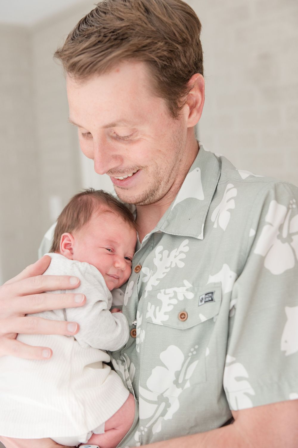 Parent wearing floral shirt tenderly holds sleeping newborn swaddled in white.