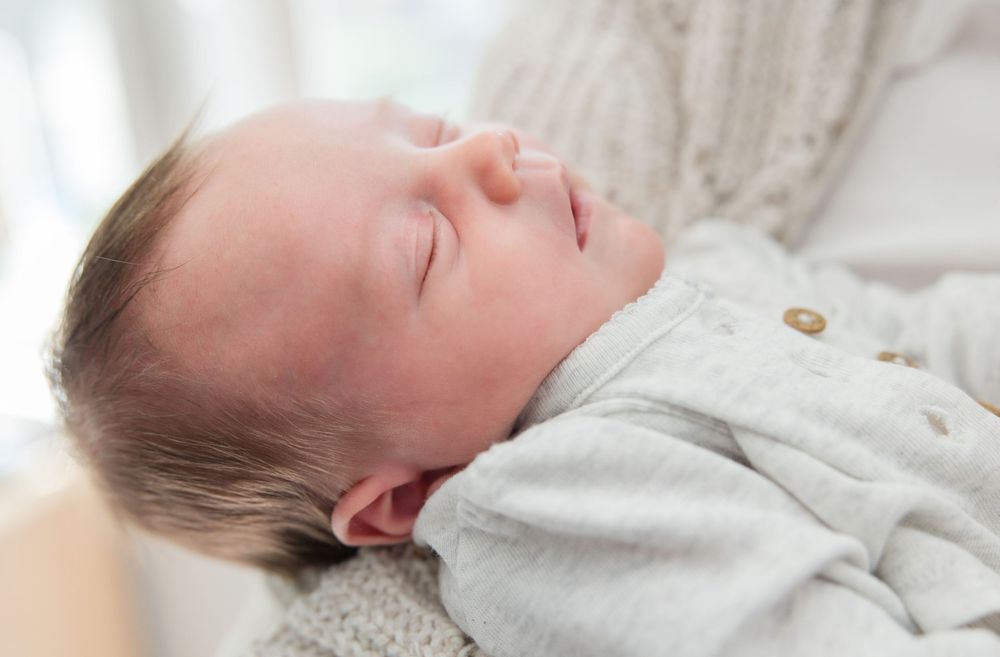 A sleeping infant wearing a light colored outfit rests peacefully on soft bedding.