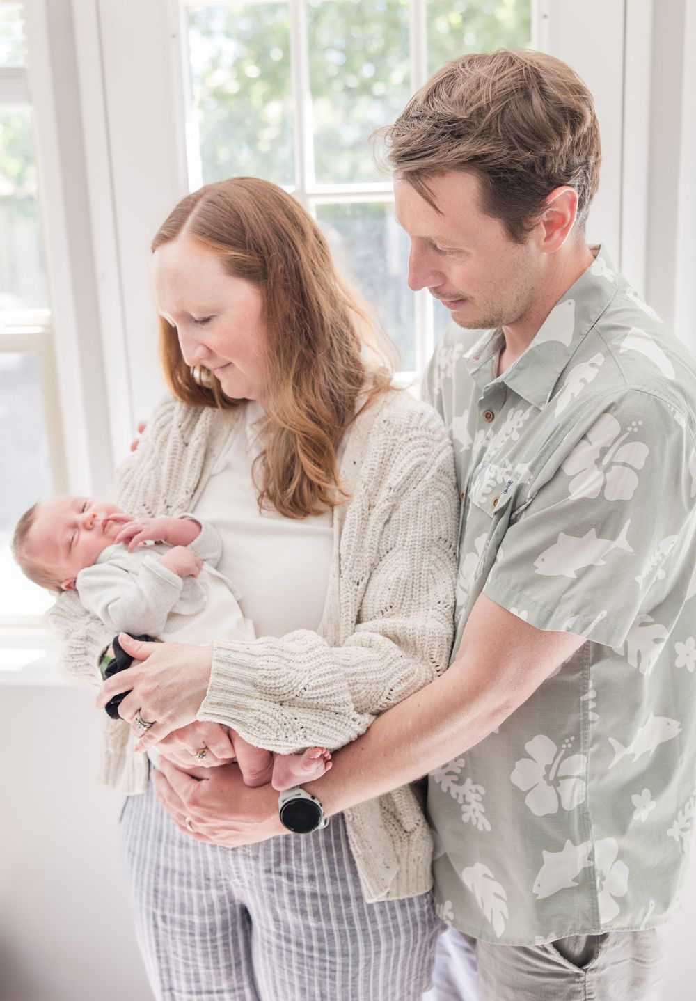 Parents in casual attire lovingly cradle their newborn baby in a bright sunlit room.
