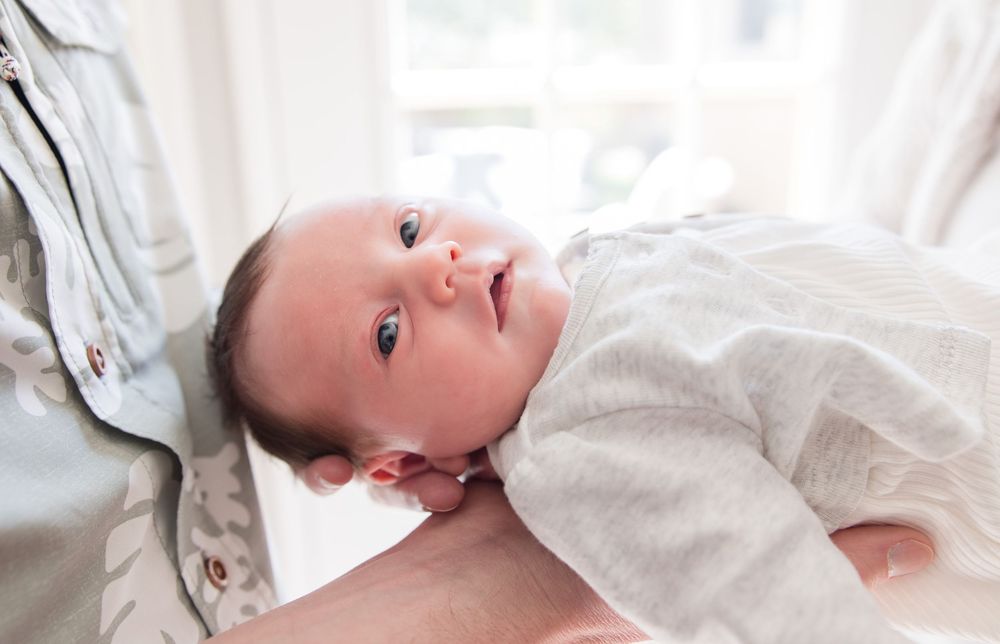 A newborn baby wearing white clothing rests on a light-colored blanket while gazing at the camera.