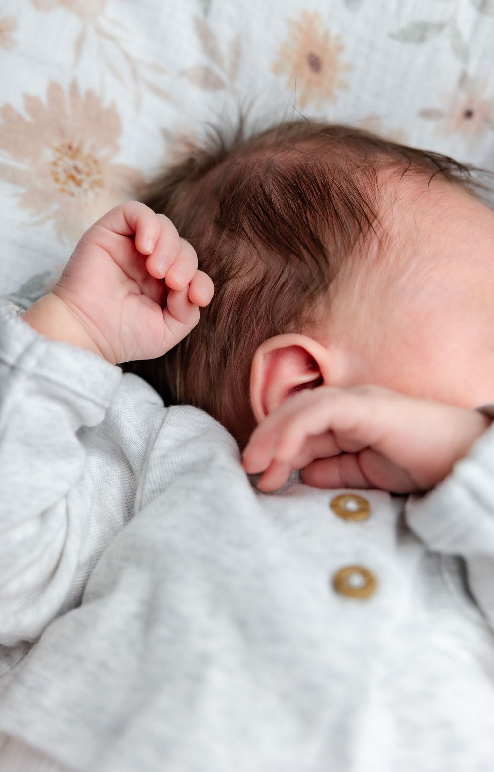 Sleeping newborn wearing white clothing with wooden buttons rests on floral patterned bedding.