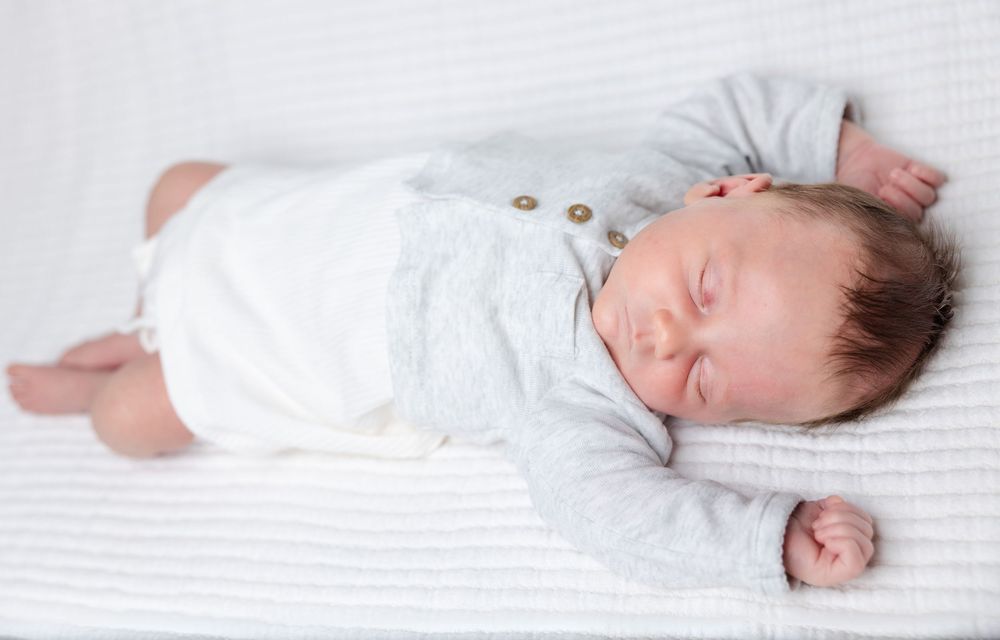 Peaceful sleeping baby in white onesie lies on soft gray blanket.