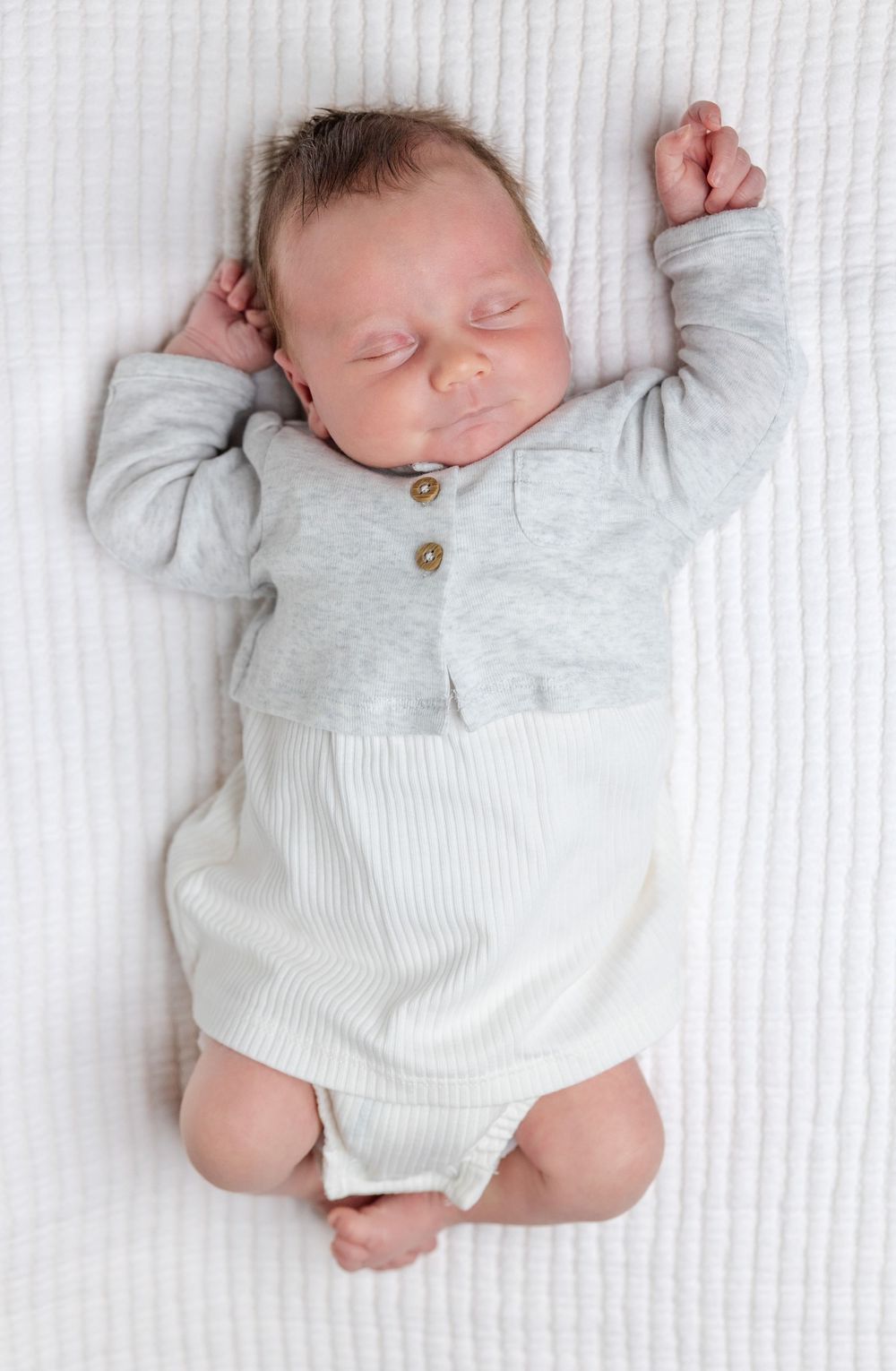 Sleeping newborn infant wearing a gray and white two-piece outfit lies on white textured fabric.