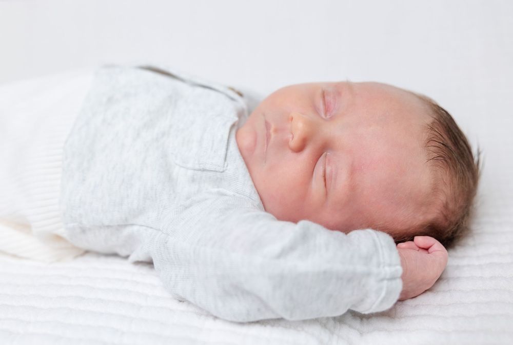 Sleeping infant wearing white clothing rests on light colored blanket.