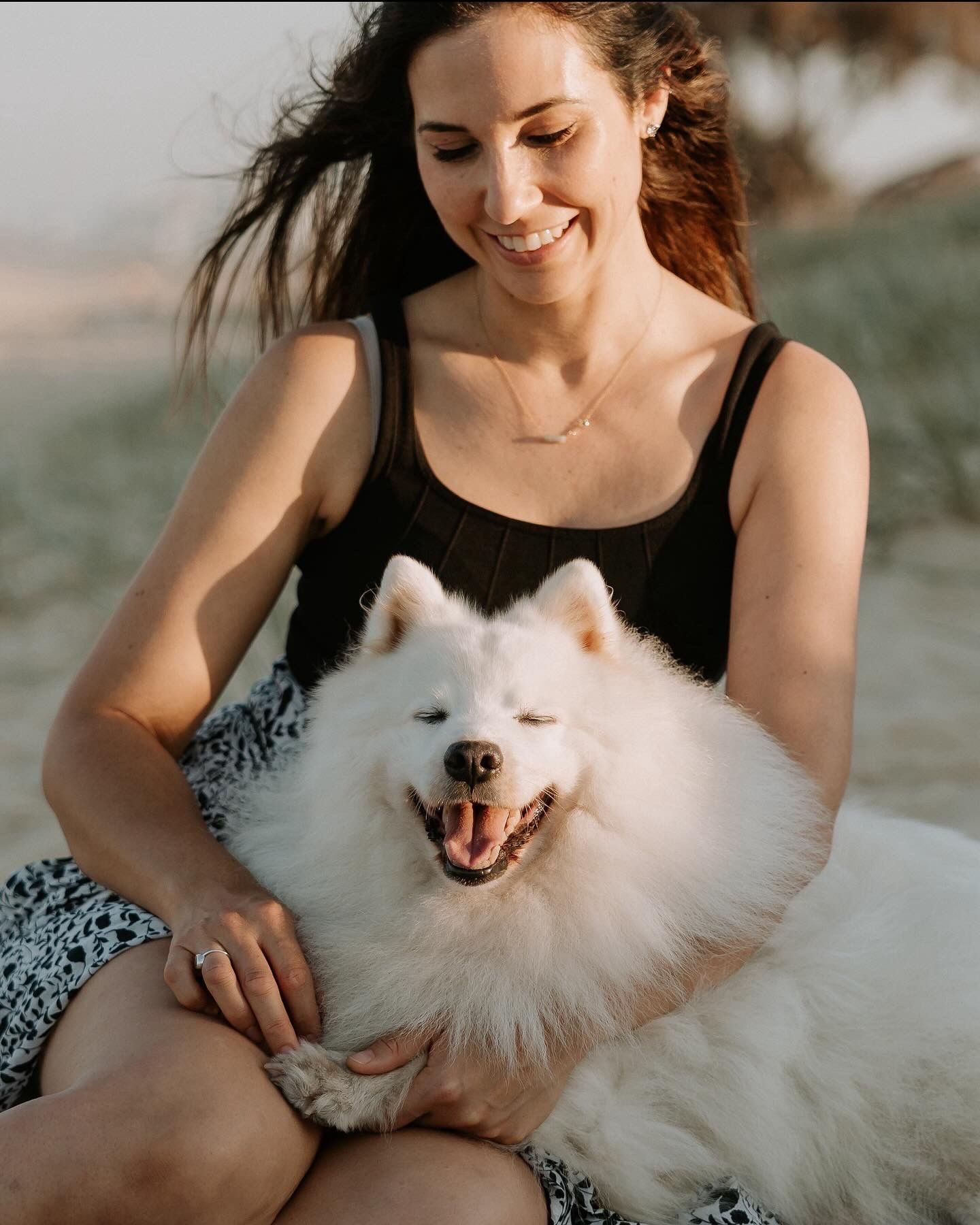 A person in black sits on the ground cuddling a fluffy white Samoyed dog with a happy expression.