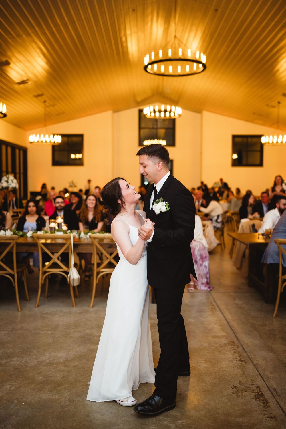 A couple shares their first dance in an elegant reception venue with warm lighting and wooden crossback chairs.