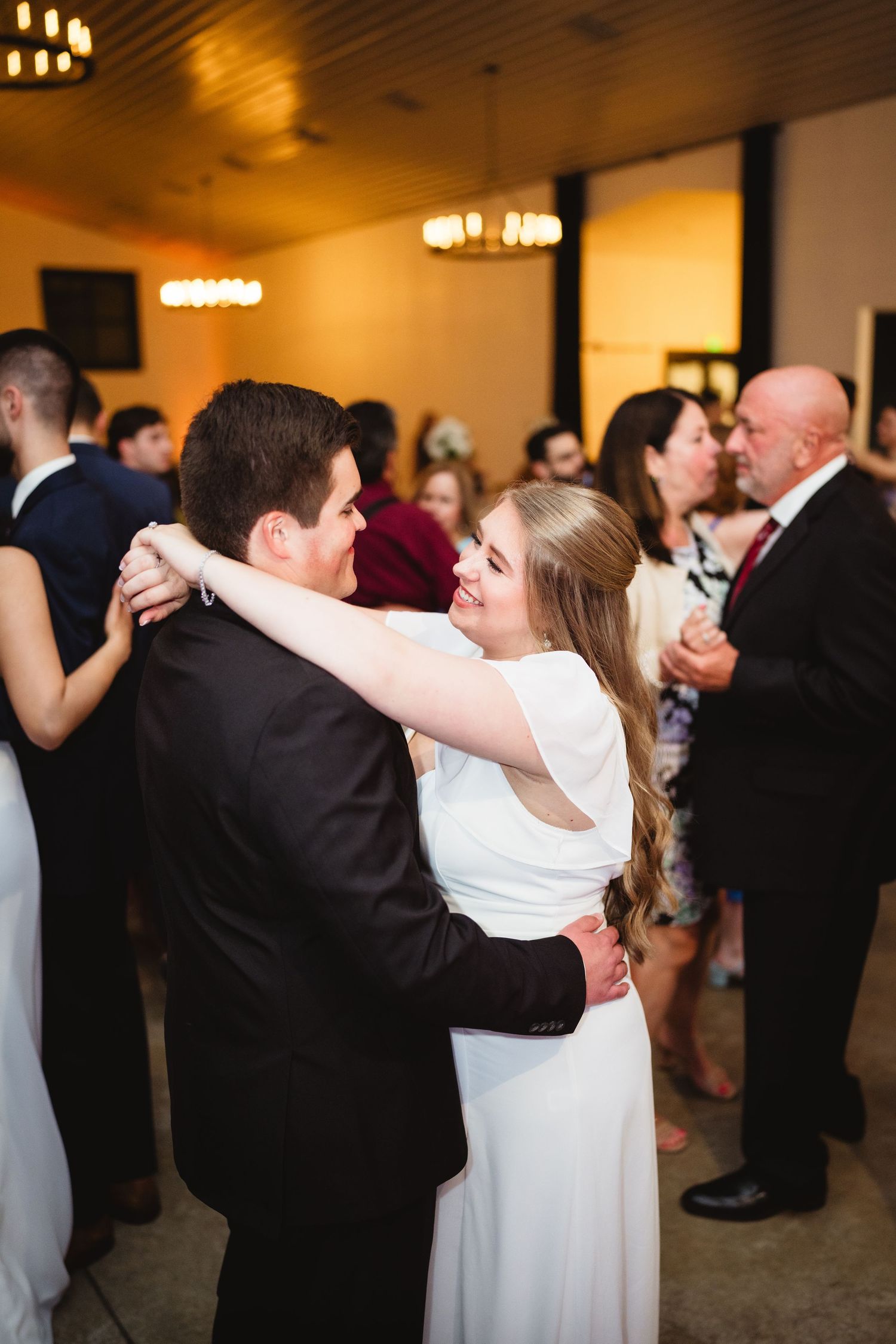 A couple shares a romantic dance together at their wedding reception while other guests dance nearby in a warmly lit venue.