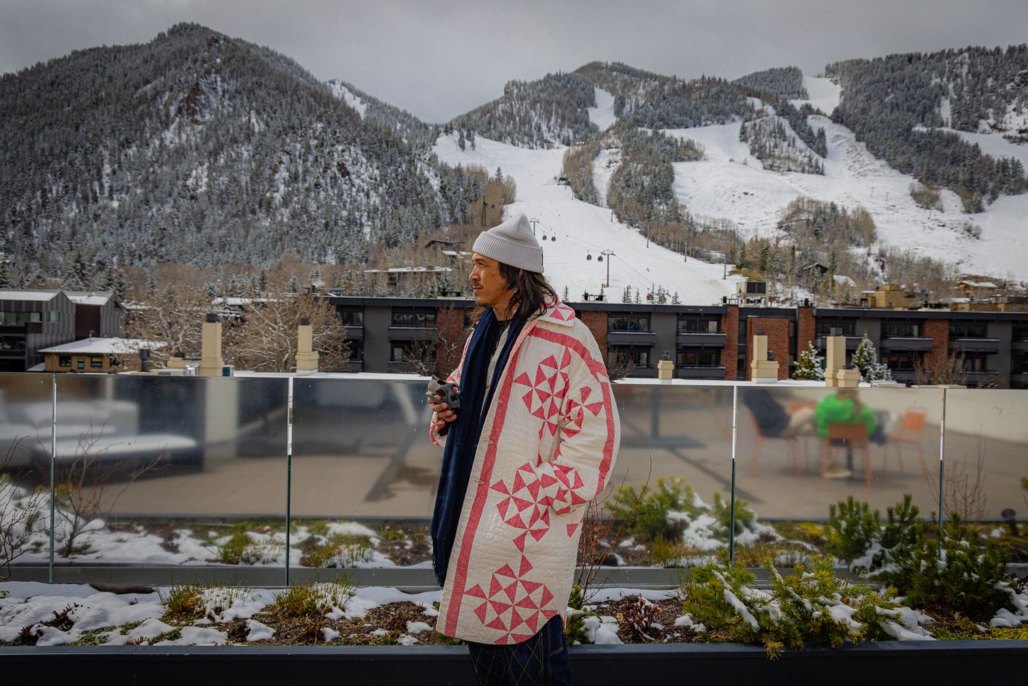 A snowboard leans against a balcony railing with snowy ski slopes and mountain resort buildings in the background.