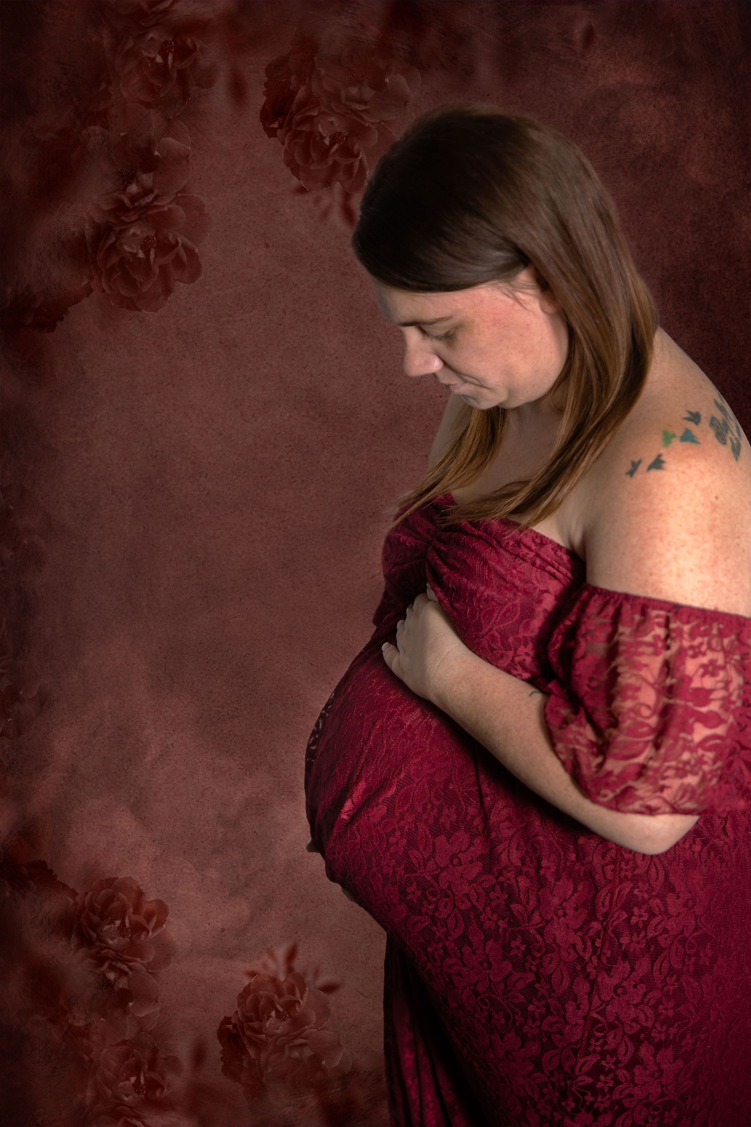 Side view maternity portrait with burgundy lace dress against moody floral backdrop.