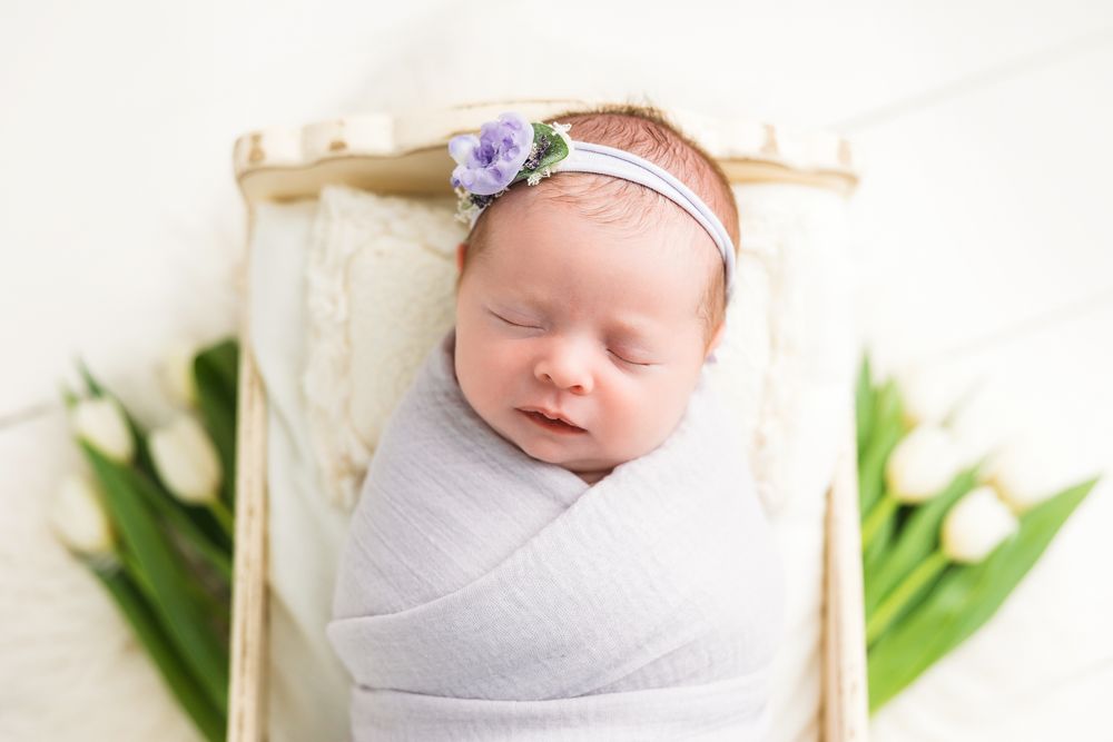 Sleeping newborn in a white wrap with purple flower headband rests in wooden crate with tulips.