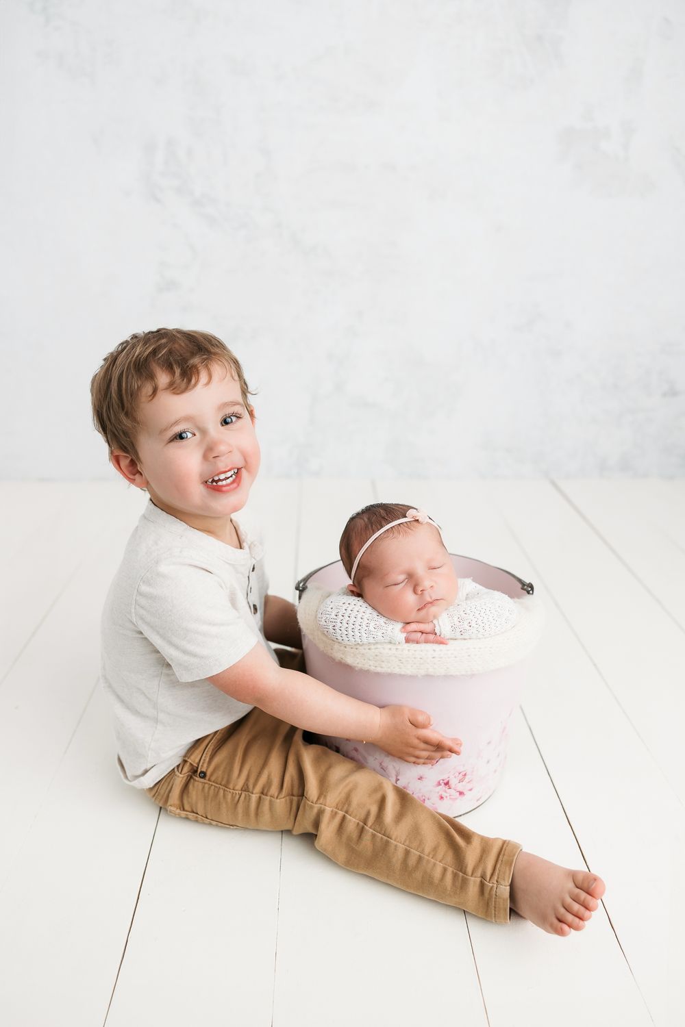 A sibling photographed in a white studio setting tenderly poses with a sleeping newborn baby.