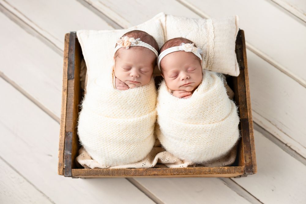 Twin newborns swaddled in cream blankets sleeping peacefully in a rustic wooden basket.