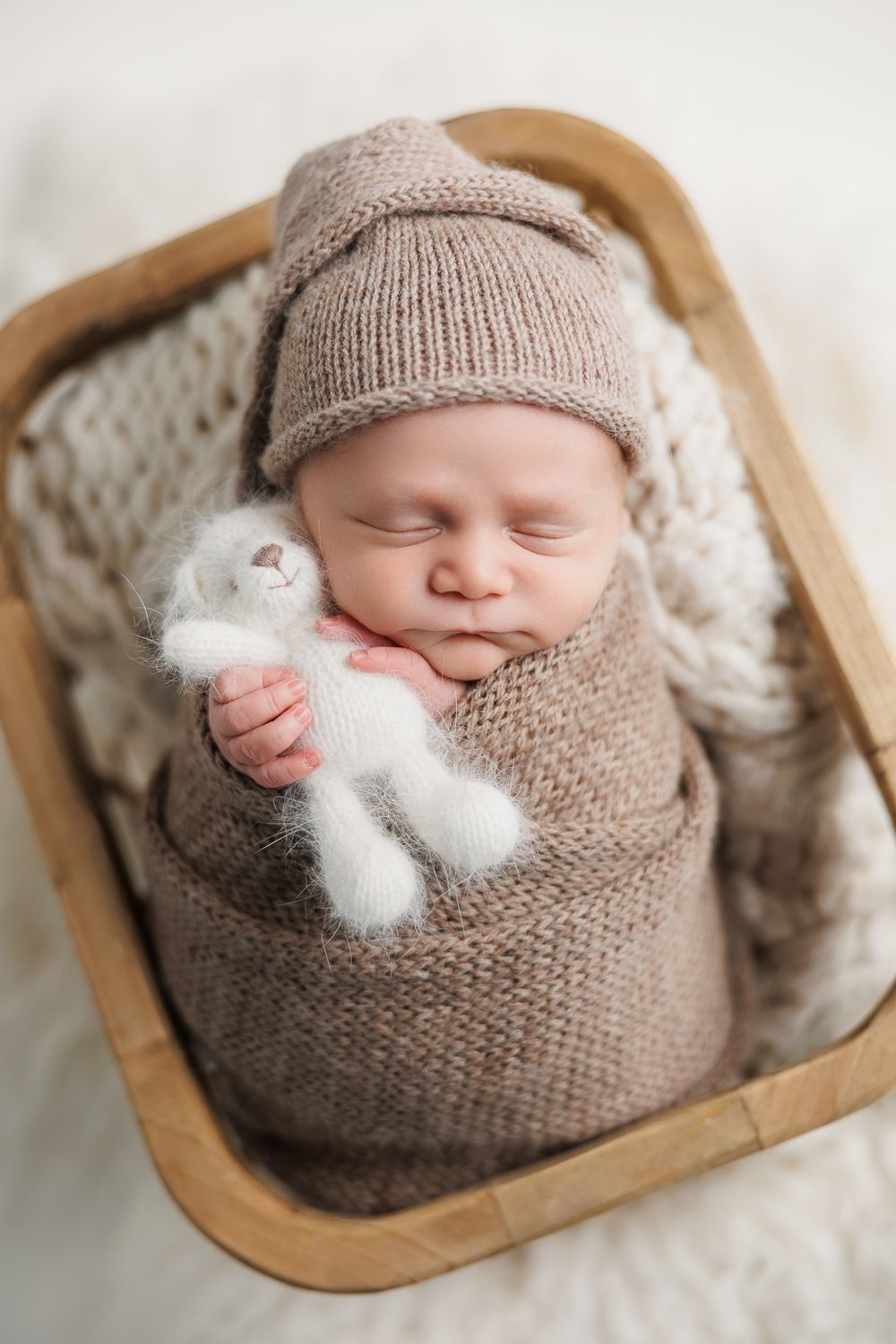 Sleeping newborn swaddled in knit beige wrap holding small white plush toy rests in wooden bowl on textured blanket.