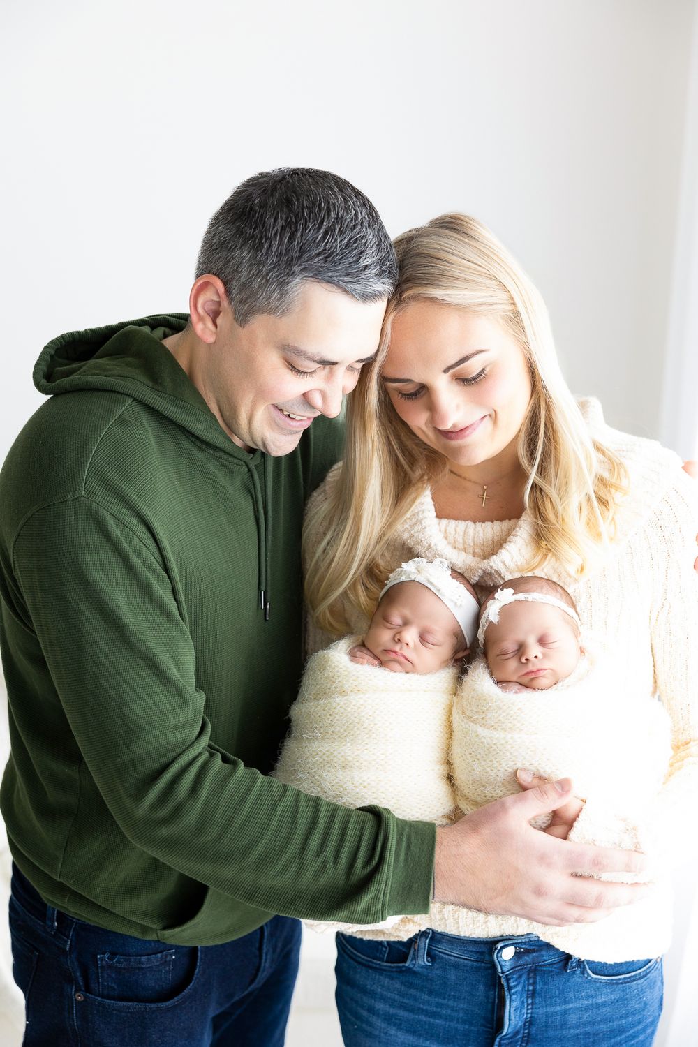 Parents in casual attire hold their sleeping newborn twins wrapped in white blankets against a light background.