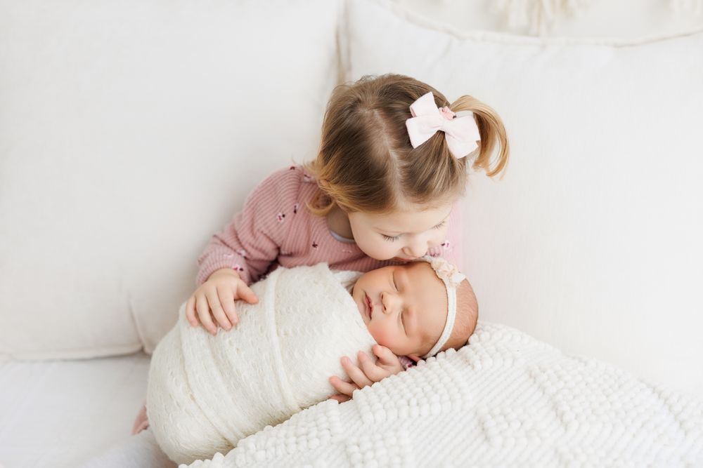 Two siblings share a tender moment on white bedding as one gently leans over the other swaddled in white.