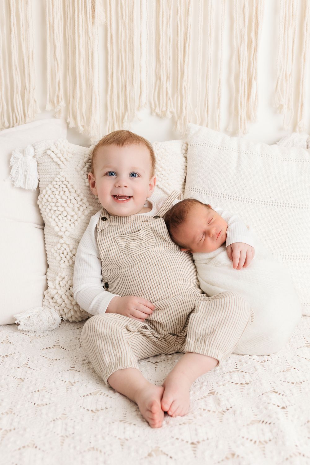 Two young children in neutral colored outfits sit together on a white knit blanket against a cream colored backdrop.
