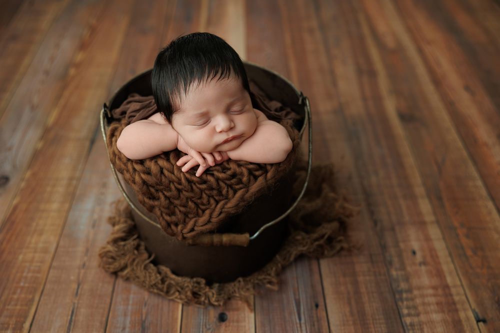 Peaceful newborn sleeping in rustic wooden container with brown textured fabric on wooden floor.