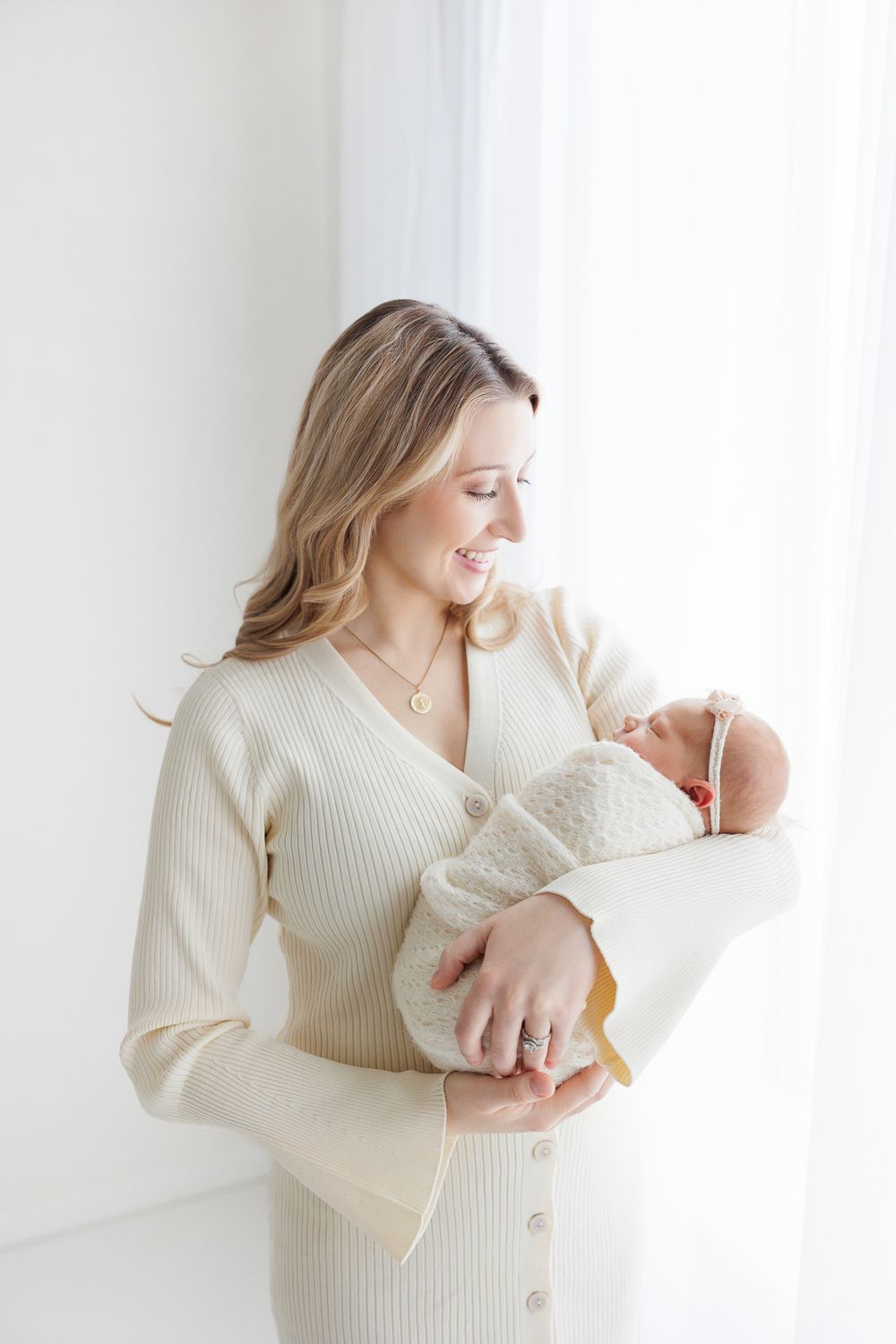 A tender moment between parent and newborn baby in a cozy white knit dress by a bright window.