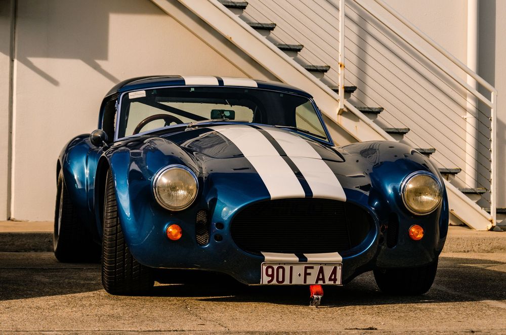 Vintage Shelby Cobra replica in blue with white racing stripes parked outside.