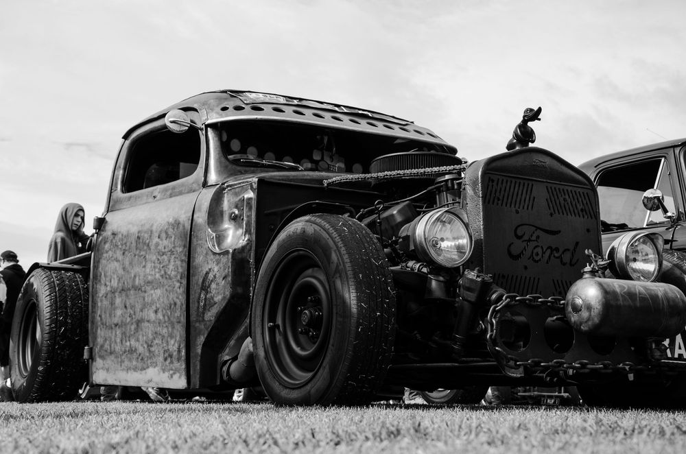 A vintage rat rod car with patina and lowered suspension parked on grass in black and white.