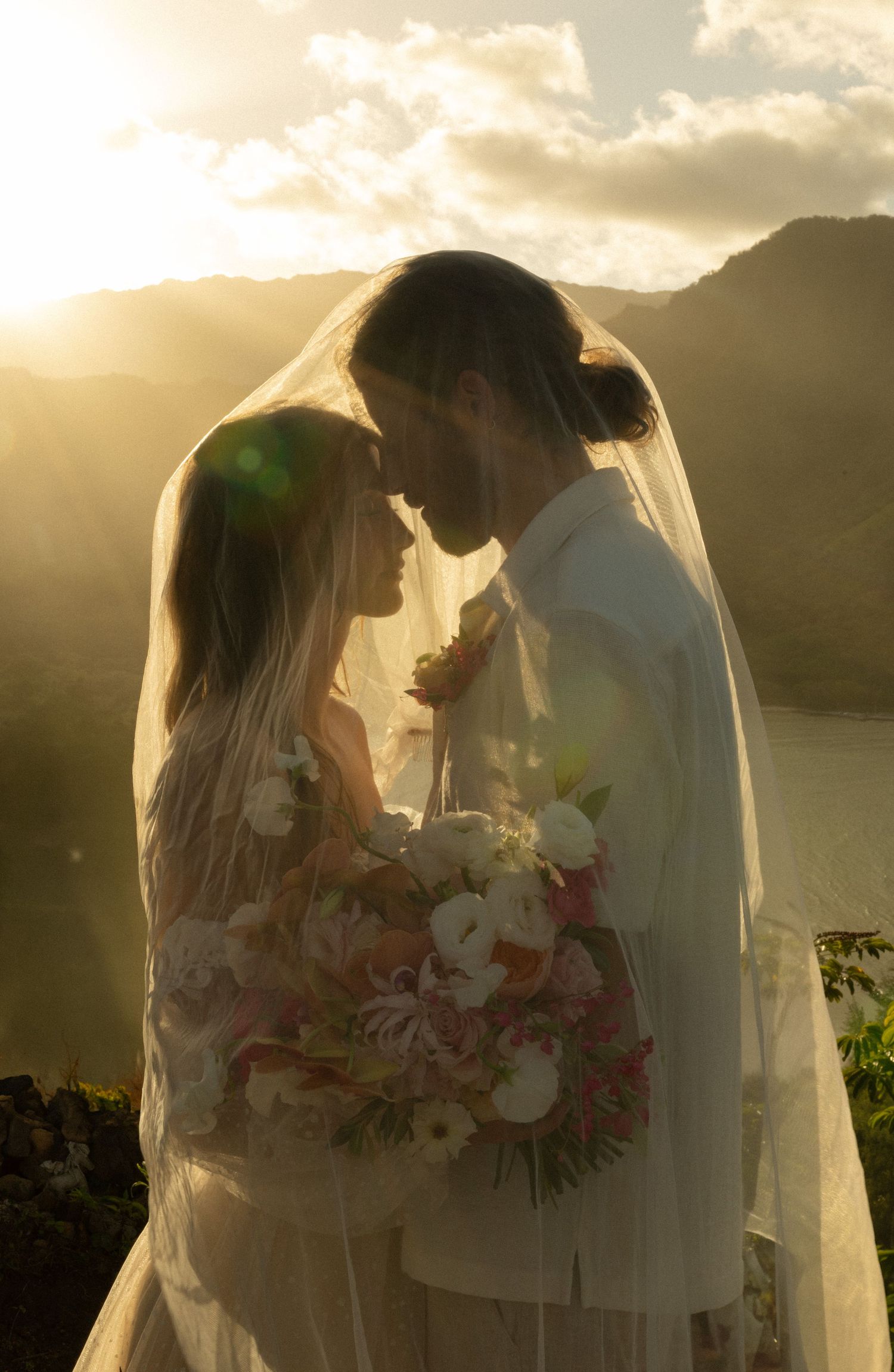 Silhouetted couple sharing an intimate moment at sunset with mountains and flowing veil, holding a pink and white bouquet.