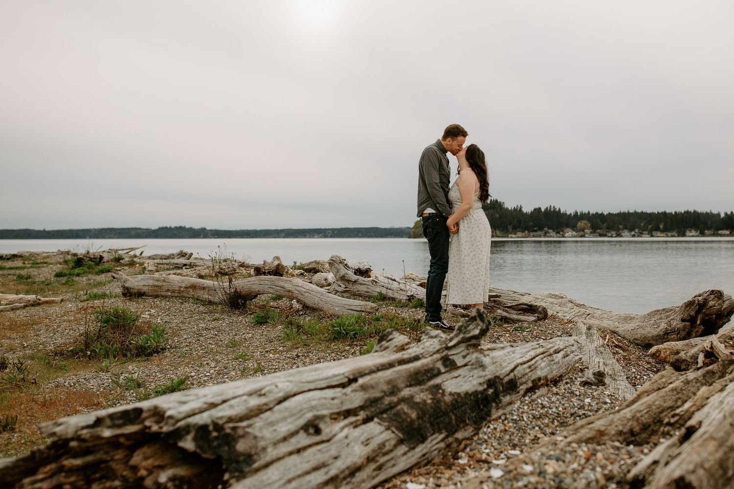 A romantic moment shared on fallen logs at the edge of a moody waterfront landscape.