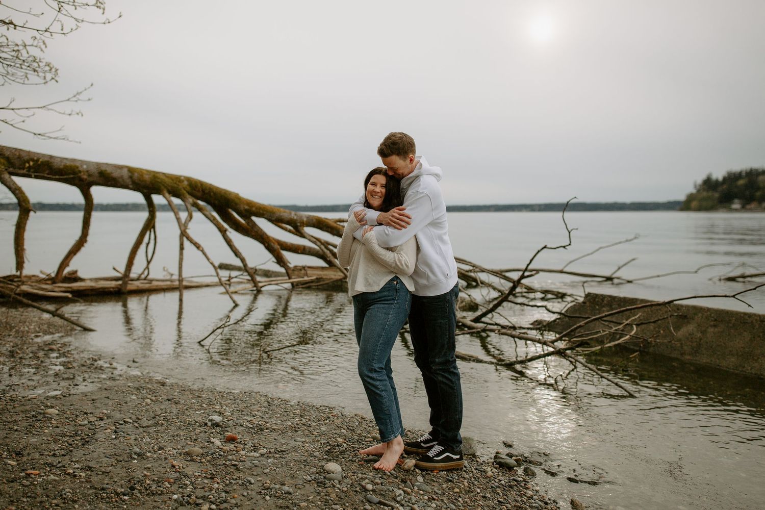 A couple embraces on a beach shoreline at sunset with driftwood and calm waters in the background.