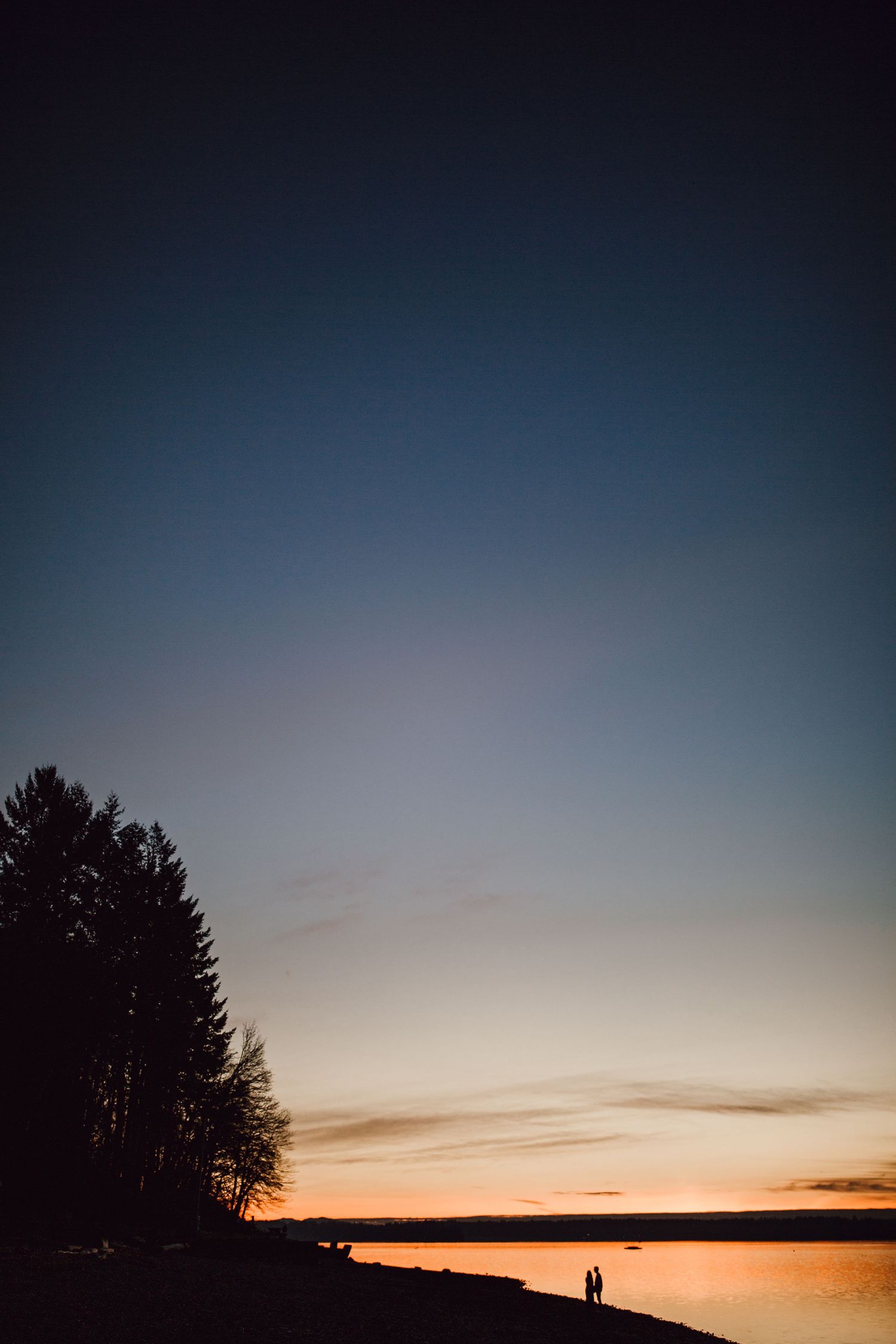 Two silhouettes stand together on a lakeside beach at dusk with evergreen trees and a dark blue gradient sky.