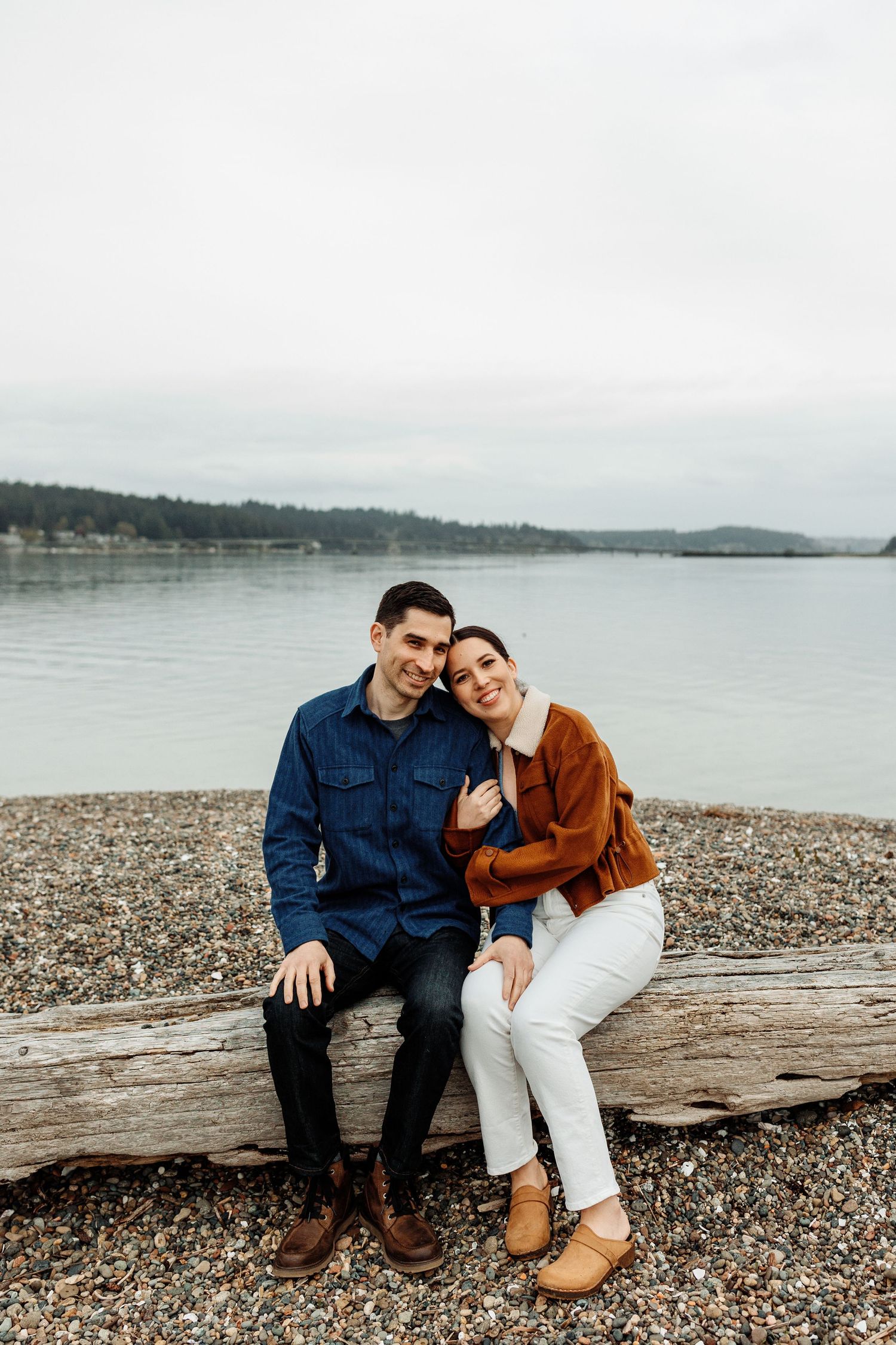 A couple sits together on driftwood at a rocky beach overlooking a serene lake on an overcast day.