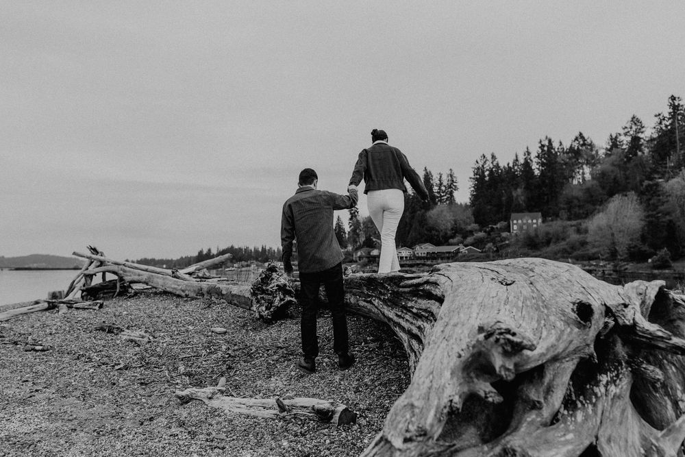 Two people explore a rocky beach with driftwood along the coastline in black and white.