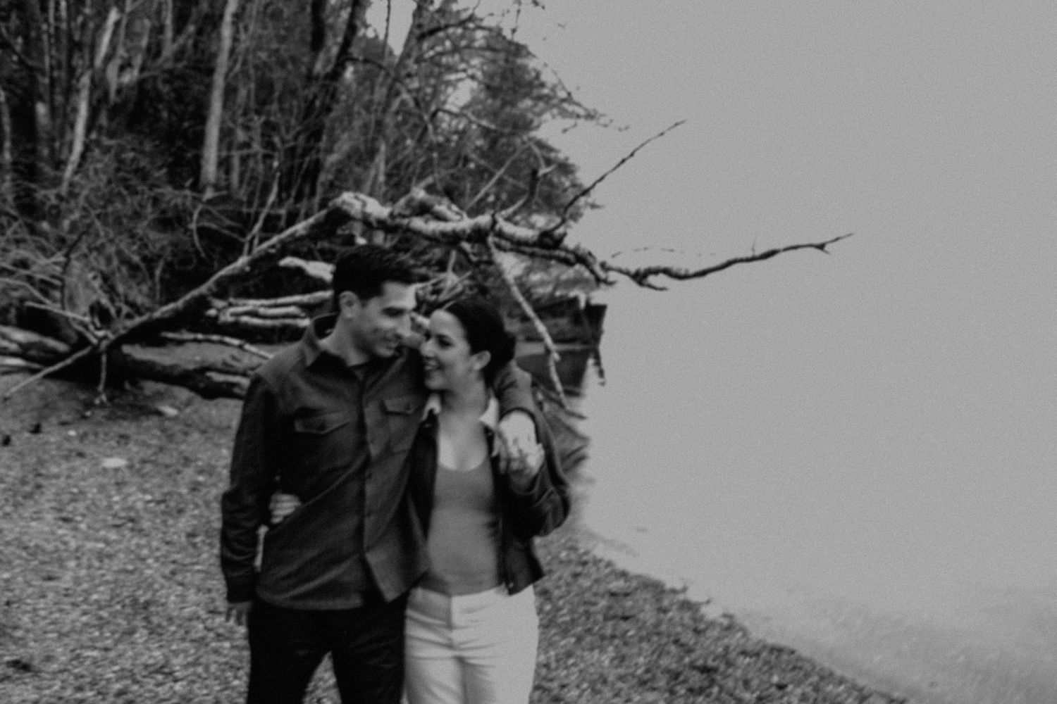 A couple stands on a forest path surrounded by bare trees and foggy skies in black and white photography.