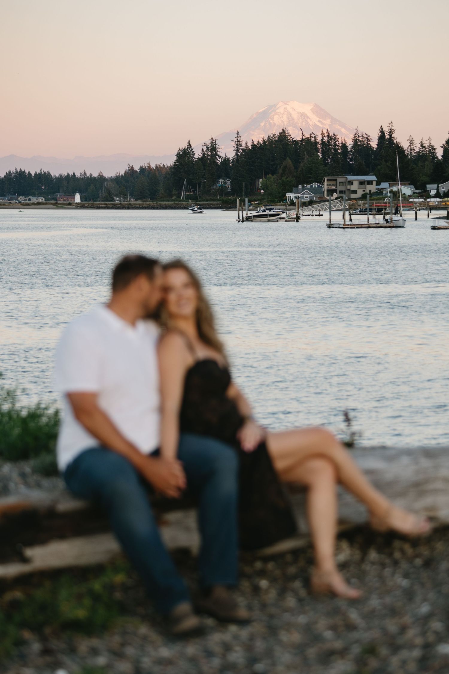 A couple sits on a rocky shore at sunset with a marina and snow-capped mountain visible in the background.