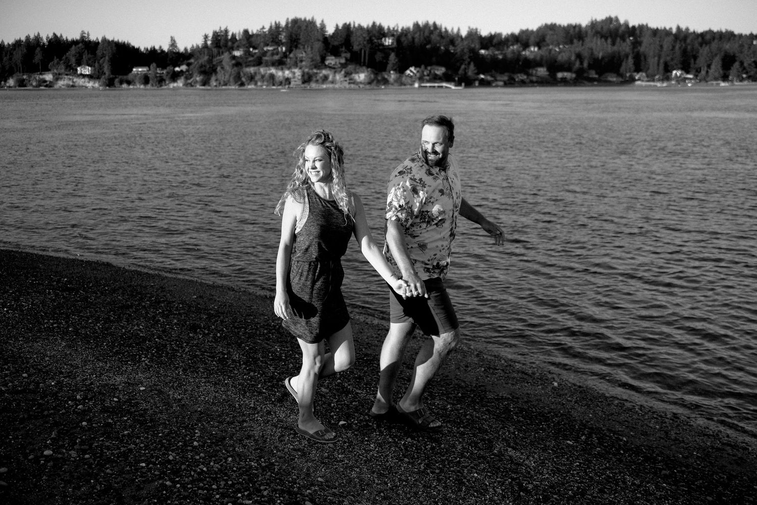 Two people hold hands while splashing joyfully in shallow water along a lakeside with tree-lined shore in background.